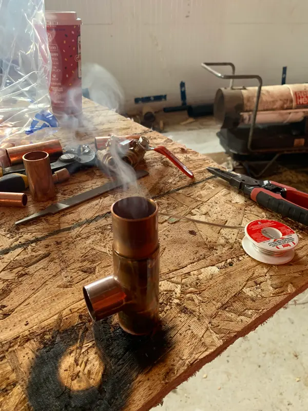 A freshly soldered copper tee standing on a plywood workbench, smoke still rising from it, with a spool of solder beside it.