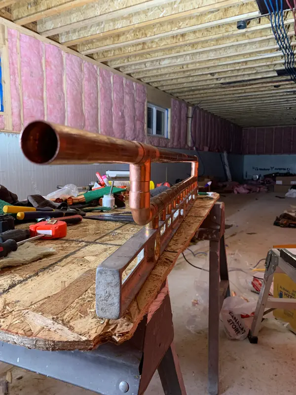 Looking down the open end of the assembled copper manifold as it rests against a long level on the bench, with the unfinished basement stretching behind.