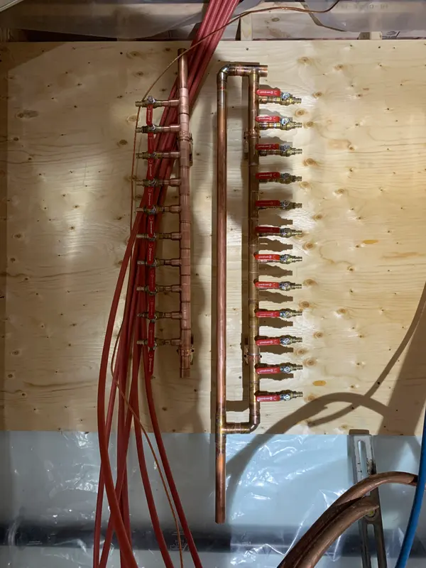 Two finished copper manifolds with red-handled valves mounted on a plywood panel, red supply lines hanging loose beside them.